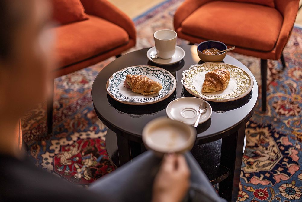 A woman sits in the Hotel Amadeus lounge with a coffee in her hand and croissants on the table